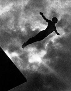 A black and white photo of a black figure in a diving pose against a background of moody clouds